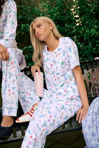 A woman with long blonde hair sits thoughtfully on an outdoor bench, surrounded by green foliage, wearing Roller Rabbit’s Game Day Evie Pajamas in a pastel print and holding a polka dot cup.