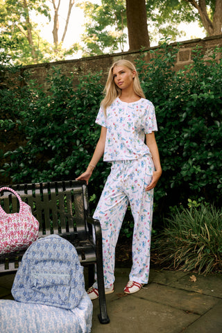 A woman with long blonde hair sits thoughtfully on an outdoor bench, surrounded by green foliage, wearing Roller Rabbit’s Game Day Evie Pajamas in a pastel print and holding a polka dot cup.