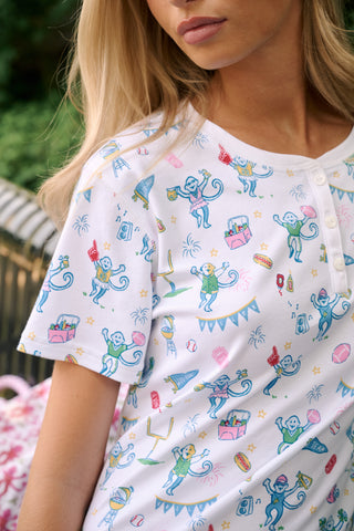 A woman with long blonde hair sits thoughtfully on an outdoor bench, surrounded by green foliage, wearing Roller Rabbit’s Game Day Evie Pajamas in a pastel print and holding a polka dot cup.