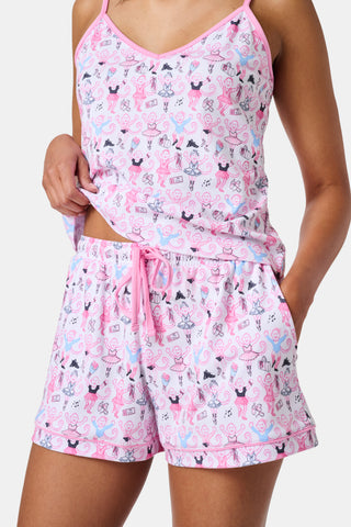A woman wearing the Roller Rabbit Monkey Pirouette Mischa Set—pink and white cat-print Pima cotton loungewear—and holding a matching bag stands in front of white lockers, smiling slightly at the camera.