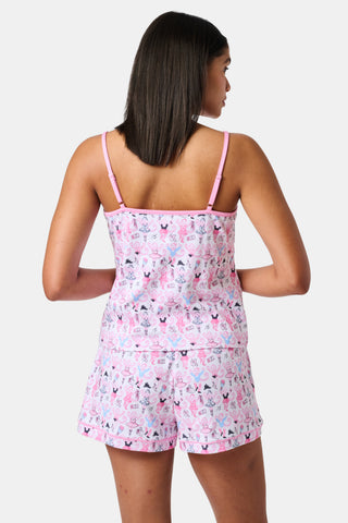 A woman wearing the Roller Rabbit Monkey Pirouette Mischa Set—pink and white cat-print Pima cotton loungewear—and holding a matching bag stands in front of white lockers, smiling slightly at the camera.