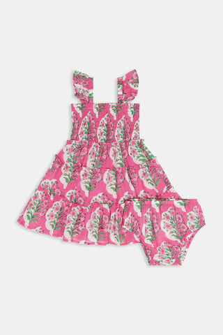 The Kids Ava Floral Ceri Dress by Roller Rabbit is a sleeveless baby dress featuring a smocked bodice and ruffled straps, adorned with green and white leaf patterns on a vibrant pink background, and comes with matching bloomers.
