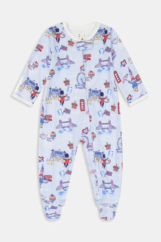 A young child with curly hair stands barefoot against a plain white background, wearing Roller Rabbit’s Kids London Monkey Pajamas—light blue long-sleeve pajamas adorned with playful, colorful patterns featuring guards, clocks, and iconic British landmarks.