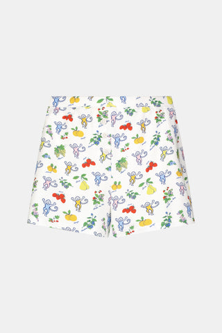 A person wearing a white sleeveless top and Roller Rabbit Monkey Fruits D'Ete Jones Shortie, featuring colorful prints of fruits, flowers, and playful cartoon figures; hands resting on the sides of the shorts.