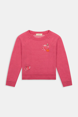 A young girl smiles, wearing the Roller Rabbit Kids Autumn Hearts Crewneck—a pink sweatshirt featuring small embroidered butterflies—paired with a white skirt adorned with a colorful heart pattern, as she stands with her hands behind her back.