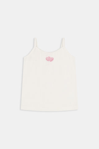 The Kids Hearts Pointelle Bridget Cami by Roller Rabbit is a white sleeveless camisole with thin straps and a small pink heart-shaped patch at the center of the chest. Crafted from soft pointelle cotton, it features a delicate scoopneck design. The background is light gray.