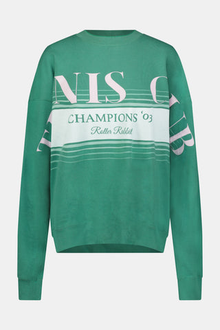 A person wearing a preppy green Roller Rabbit Tennis Champions Andie Sweatshirt stands in front of bookshelves. Only the lower part of their face is visible, and their hands are in their pockets.