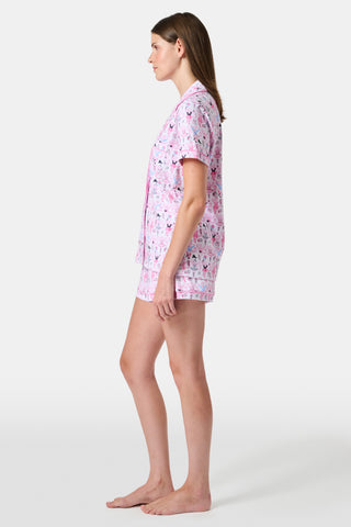 The Monkey Pirouette Polo Pajamas by Roller Rabbit feature a pink short-sleeve button-up top and matching shorts, adorned with a colorful and playful monkey, animal, and floral pattern on a white background.