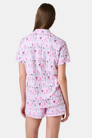 The Monkey Pirouette Polo Pajamas by Roller Rabbit feature a pink short-sleeve button-up top and matching shorts, adorned with a colorful and playful monkey, animal, and floral pattern on a white background.