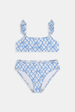 A young girl with curly hair smiles in the Roller Rabbit Kids Seaside Knots Rory Bikini, featuring ruffled shoulder straps and UPF 50+ sun protection, posing against a plain light background.