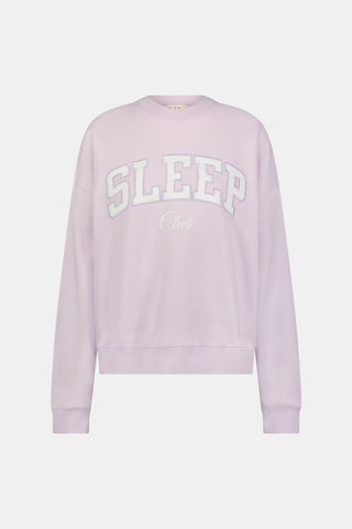 A woman wearing the Roller Rabbit Sleep Club Andie Sweatshirt in pastel lavender, paired with patterned white shorts, stands and smiles against a plain white background.