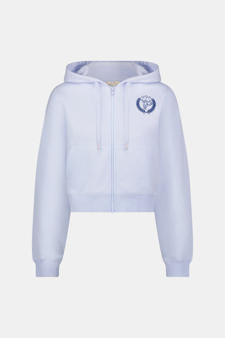 A woman wears the Roller Rabbit RR Athletic Club Cropped Zip Up—a pastel blue terry hoodie with an athletic club logo on the chest—paired with a blue-and-white checkered skirt, standing against a white background.