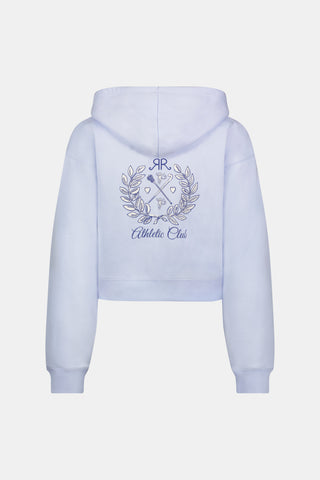 A woman wears the Roller Rabbit RR Athletic Club Cropped Zip Up—a pastel blue terry hoodie with an athletic club logo on the chest—paired with a blue-and-white checkered skirt, standing against a white background.