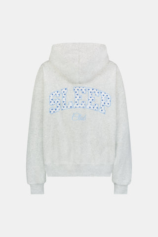 A person is wearing the Roller Rabbit Sleep Club Mimi Sweatpants in light gray, featuring wide legs and crafted from velvety soft Dream Fleece with “Sleep Club” printed in light blue on the left thigh, paired with white sneakers and a cropped white top. The background is plain white.