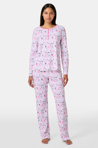 A woman with long brown hair stands next to white lockers, looking at the camera and holding a box, wearing Roller Rabbit's Monkey Pirouette Pajamas featuring a pink-patterned dancing monkey print in soft Pima cotton.