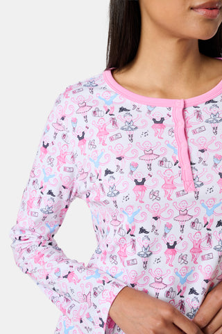 A woman with long brown hair stands next to white lockers, looking at the camera and holding a box, wearing Roller Rabbit's Monkey Pirouette Pajamas featuring a pink-patterned dancing monkey print in soft Pima cotton.