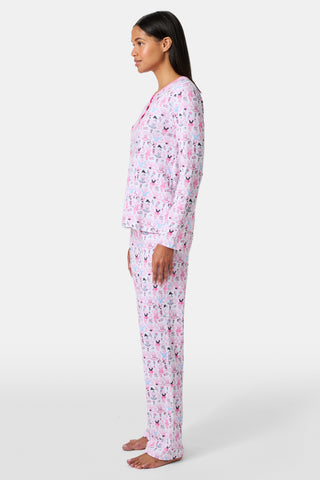 A woman with long brown hair stands next to white lockers, looking at the camera and holding a box, wearing Roller Rabbit's Monkey Pirouette Pajamas featuring a pink-patterned dancing monkey print in soft Pima cotton.