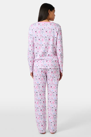 A woman with long brown hair stands next to white lockers, looking at the camera and holding a box, wearing Roller Rabbit's Monkey Pirouette Pajamas featuring a pink-patterned dancing monkey print in soft Pima cotton.