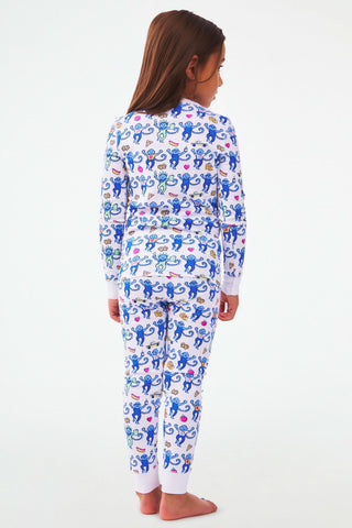 A young child with long brown hair stands barefoot, facing away in a plain, light-colored studio, wearing Kids NY Monkey Pajamas—matching Pima cotton sleepwear with a playful animal print on a white background.