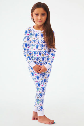 A young girl stands barefoot against a white background, wearing Kids NY Monkey Pajamas—white Pima cotton with colorful monkey print. She looks at the camera with a slight smile, her long brown hair down and hands clasped in front.