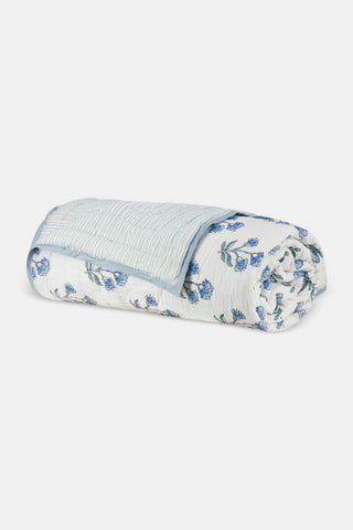 A neatly made bed with the Roller Rabbit Rosa Ditsy Floral Quilt sits beside a round wicker nightstand with a vase of white flowers and stacked books, atop a woven rug and framed by white curtains in the background.