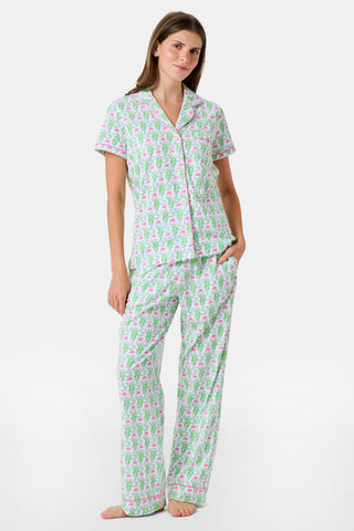 A woman stands barefoot against a plain white background, wearing the Roller Rabbit Monkey Fiora Pajamas—a matching set crafted from soft Pima cotton for extra comfort. The pajama set features a short-sleeve button-up top and long pants adorned with a pastel floral motif.
