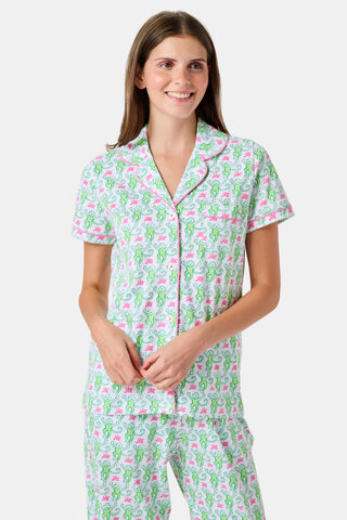 A woman stands barefoot against a plain white background, wearing the Roller Rabbit Monkey Fiora Pajamas—a matching set crafted from soft Pima cotton for extra comfort. The pajama set features a short-sleeve button-up top and long pants adorned with a pastel floral motif.