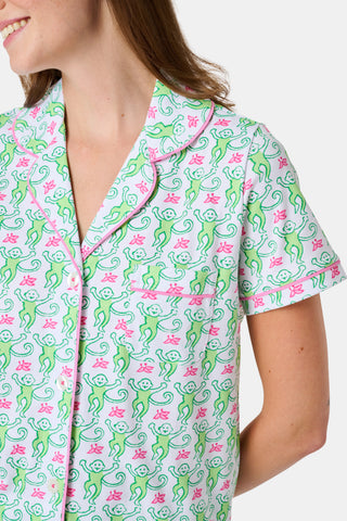 A woman stands barefoot against a plain white background, wearing the Roller Rabbit Monkey Fiora Pajamas—a matching set crafted from soft Pima cotton for extra comfort. The pajama set features a short-sleeve button-up top and long pants adorned with a pastel floral motif.