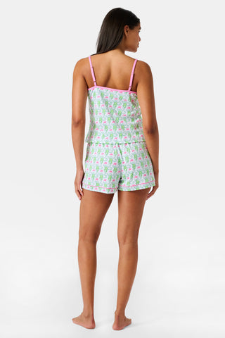 A woman wearing the SERFLEX Monkey Mischa Set, a coordinated sleeveless top and shorts with pink straps and a fun pink and green flamingo print, made from soft Pima cotton, stands against a plain white background.