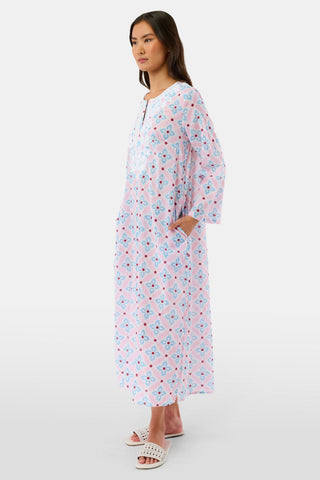 A woman with long hair wears the Roller Rabbit Lerato Inez Long Kurta, a cotton voile long sleeve pullover dress featuring a pastel pink and blue geometric pattern and white embroidery on the chest. She pairs it with white open-toe sandals.