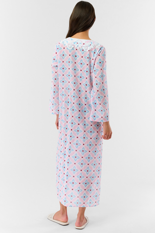 A woman with long hair wears the Roller Rabbit Lerato Inez Long Kurta, a cotton voile long sleeve pullover dress featuring a pastel pink and blue geometric pattern and white embroidery on the chest. She pairs it with white open-toe sandals.