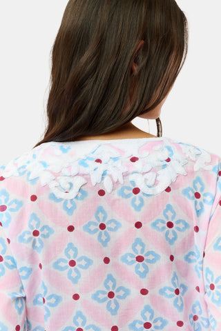 A woman with long hair wears the Roller Rabbit Lerato Inez Long Kurta, a cotton voile long sleeve pullover dress featuring a pastel pink and blue geometric pattern and white embroidery on the chest. She pairs it with white open-toe sandals.