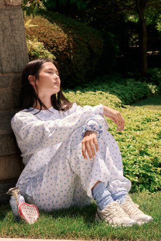 A person wearing a white Roller Rabbit Mini Monkey Blossom Andie Sweatshirt, featuring small blue and green Mini Monkey Blossom prints and large SLEEP Club lettering, stands outdoors, partially in sunlight. Their face is mostly out of frame.