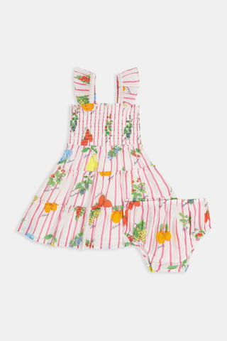 A young girl with straight brown hair wears the Roller Rabbit Kids Fruits D'Ete Ceri Dress, a pink and white smocked sundress with ruffled sleeves, and white sandals, standing and smiling against a light gray background.