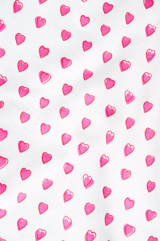 A brown poodle sits on a white background wearing Roller Rabbit's Hearts Dog Pajamas, adorable Pima Cotton pet PJs featuring small pink heart patterns for perfect matching dog pajamas.