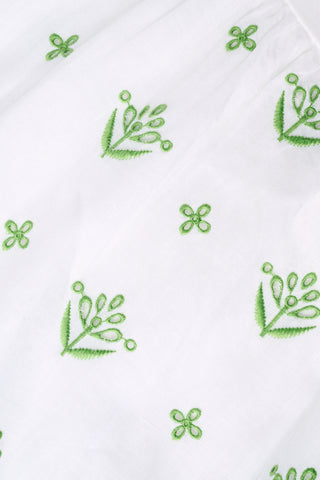 A woman with curly hair wears the Roller Rabbit Fruit Eyelet Selena Dress, a green linen maxi with floral embroidery and thin straps. She stands facing forward in yellow open-toe sandals against a plain white background.