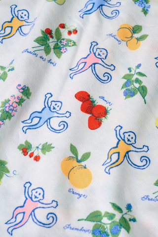 A woman wears the Monkey Fruits D’Ete Clarkson Cami by Roller Rabbit, a sleeveless white cotton-modal cami with light blue trim and a colorful fruit and cartoon print. The matching bottoms are partially visible against a plain white background.