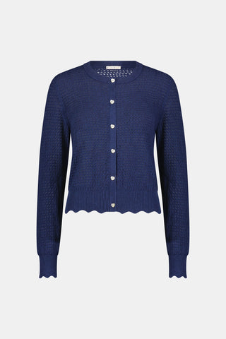 A woman wears the Roller Rabbit Pointelle Rosella Cardigan, a navy blue button-up with heart-shaped buttons, paired with white pants featuring a blue floral print, standing against a plain light background.