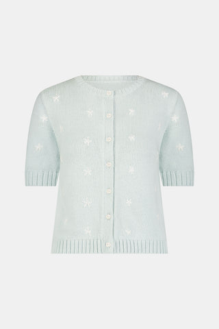 A woman with curly hair wears the Roller Rabbit Floral Bloom Nyla Cardigan, a light blue short-sleeved top with small white flowers, paired with loose white embroidered pants, standing against a plain light background.