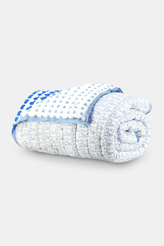 A bed features the Roller Rabbit Monkey/Hearts Reversible Quilt in light blue and white, with three matching pillows and a white headboard. Beside it, a round wicker side table holds flowers and books atop a woven rug.