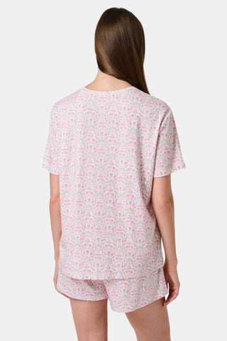 A woman wearing the Roller Rabbit Garden Gate Floral Elena Set stands by a stone wall and glass door, holding a patterned clutch and water bottle, with a matching sweatshirt tied at her waist.