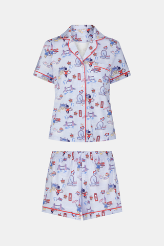 A woman stands and smiles against a plain white background, wearing the Roller Rabbit London Monkey Polo Pajamas—a light blue Pima cotton set featuring cartoon prints and red trim.