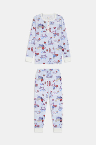 A young child with curly hair stands barefoot against a plain white background, wearing Roller Rabbit’s Kids London Monkey Pajamas—light blue long-sleeve pajamas adorned with playful, colorful patterns featuring guards, clocks, and iconic British landmarks.