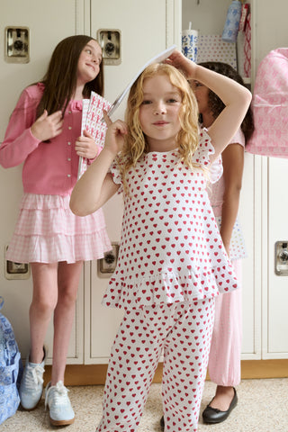 A child with long blonde hair wears the Roller Rabbit Kids Hearts Riley Set made from Pima Waffle fabric, featuring red hearts and ruffled cap sleeves, while holding a notebook with a decorative cover and standing in front of lockers.