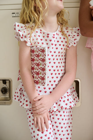 A child with long blonde hair wears the Roller Rabbit Kids Hearts Riley Set made from Pima Waffle fabric, featuring red hearts and ruffled cap sleeves, while holding a notebook with a decorative cover and standing in front of lockers.