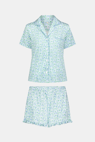 The Roller Rabbit Garden Gate Floral Wyatt Polo Pajamas feature a light blue short-sleeve set crafted from soft Pima Cotton with a delicate Garden Gate Floral Ditsy pattern. The button-up top is designed with a collar and chest pocket, while the matching shorts are accented with a charming ruffled hem. The background is plain white.
