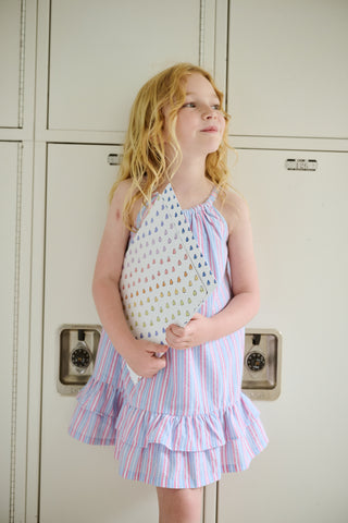 A young girl wearing the Roller Rabbit Kids Sweet Stripe Casey Dress stands in front of lockers, smiling and holding a folder decorated with colorful raindrops.