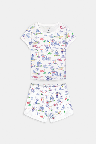 A young child stands barefoot, smiling and facing forward, wearing Roller Rabbit Kids Monkey On The Vineyard Pajamas made of soft Pima cotton with colorful cartoon patterns of animals, vehicles, and buildings. The pajamas feature buttons at the neckline.