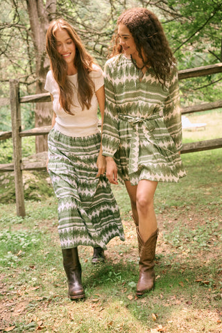 A person wearing the Roller Rabbit Hayam Carlisa Skirt in an olive and white print, paired with a cream knit top and brown boots, stands against a plain white background.