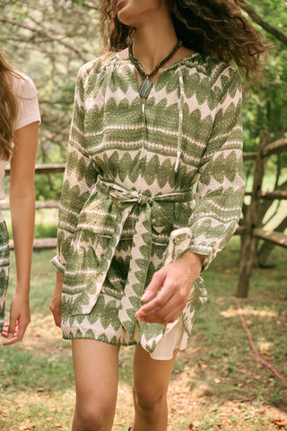 A woman outdoors wears the Hayam Monroe Dress by Roller Rabbit—an olive and white long-sleeved dress with a matching belt. She walks with another person, with trees and a wooden fence in the background.